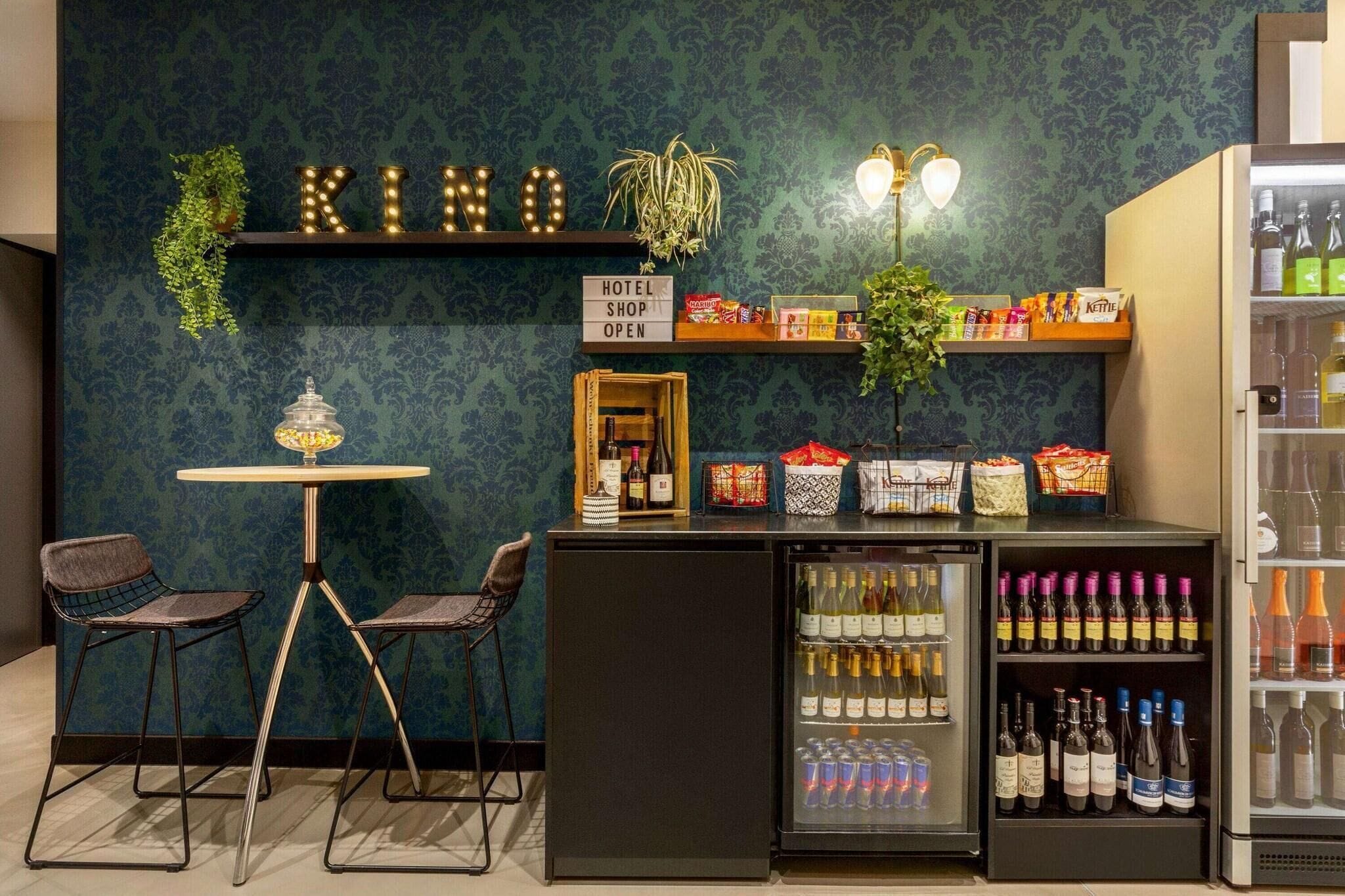 Zona de bar con un cartel que indica que la tienda del hotel está abierta, un refrigerador lleno de bebidas y un área de estar con sillas.