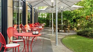 An outdoor seating area with red chairs and white umbrellas, surrounded by greenery. - City Hotel MAX - Munich West (Munich)