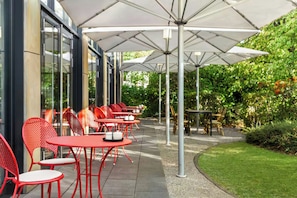 An outdoor seating area with red chairs and white umbrellas, surrounded by greenery. - City Hotel MAX - Munich West (Munich)