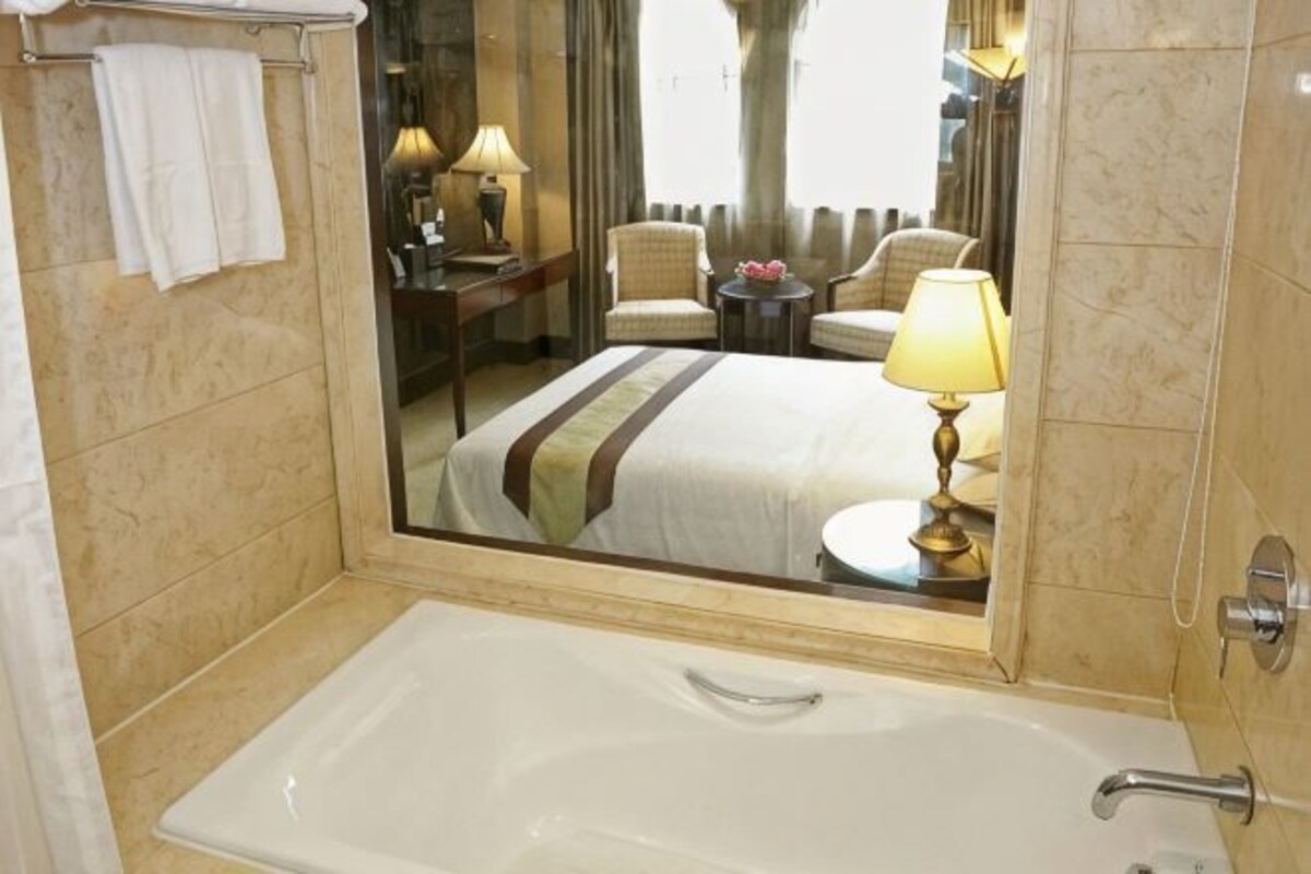 A hotel bathroom with a large mirror, a bathtub, a bed with a striped bedspread, a bedside table with a lamp, and a chair.