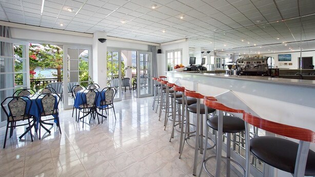 A modern bar area with high stools and a long counter, surrounded by large windows offering a view of greenery. - Athina (Kefalonia)