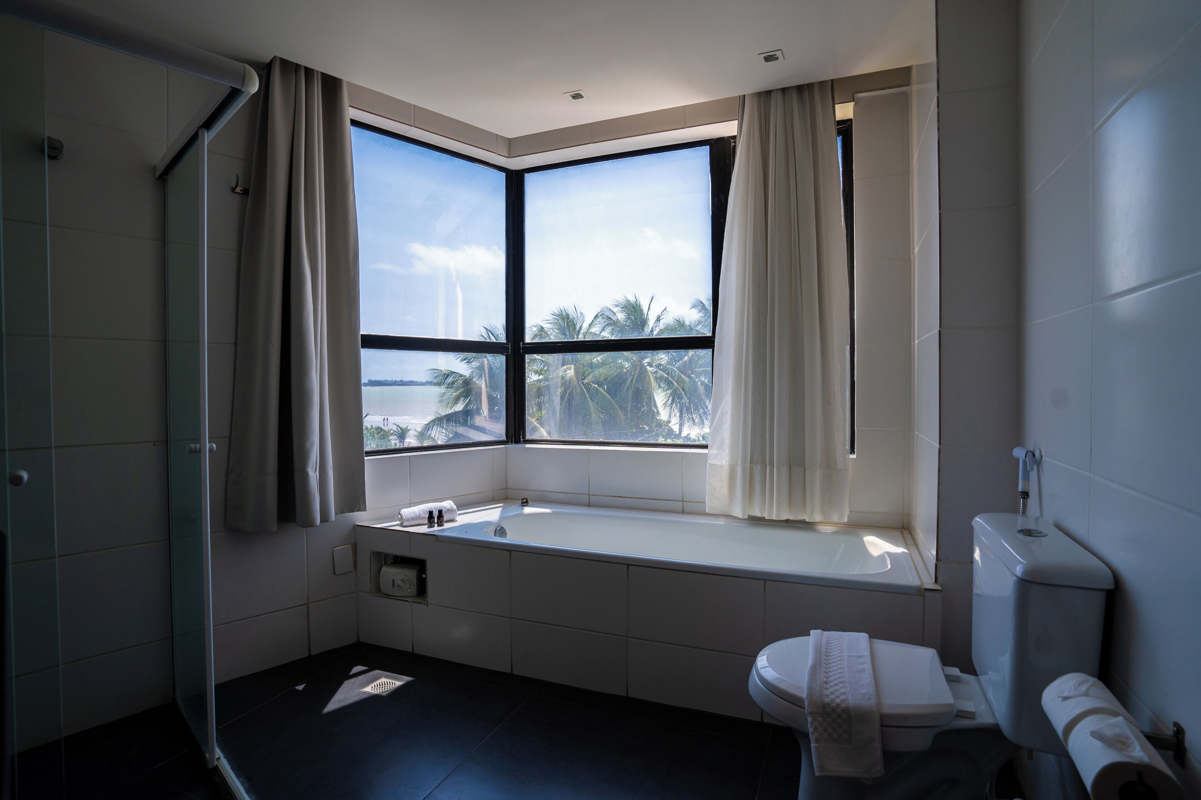 A modern bathroom with a bathtub, toilet, and large windows overlooking palm trees.