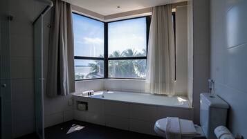 A modern bathroom with a bathtub, toilet, and large windows overlooking palm trees.
