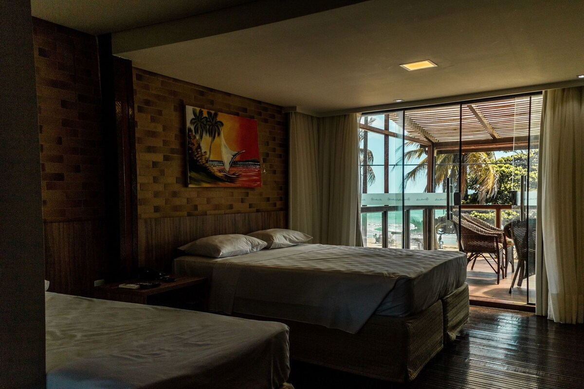 Habitación de hotel con dos camas, un cuadro en la pared, una ventana con vistas a palmeras y un balcón con muebles de mimbre.