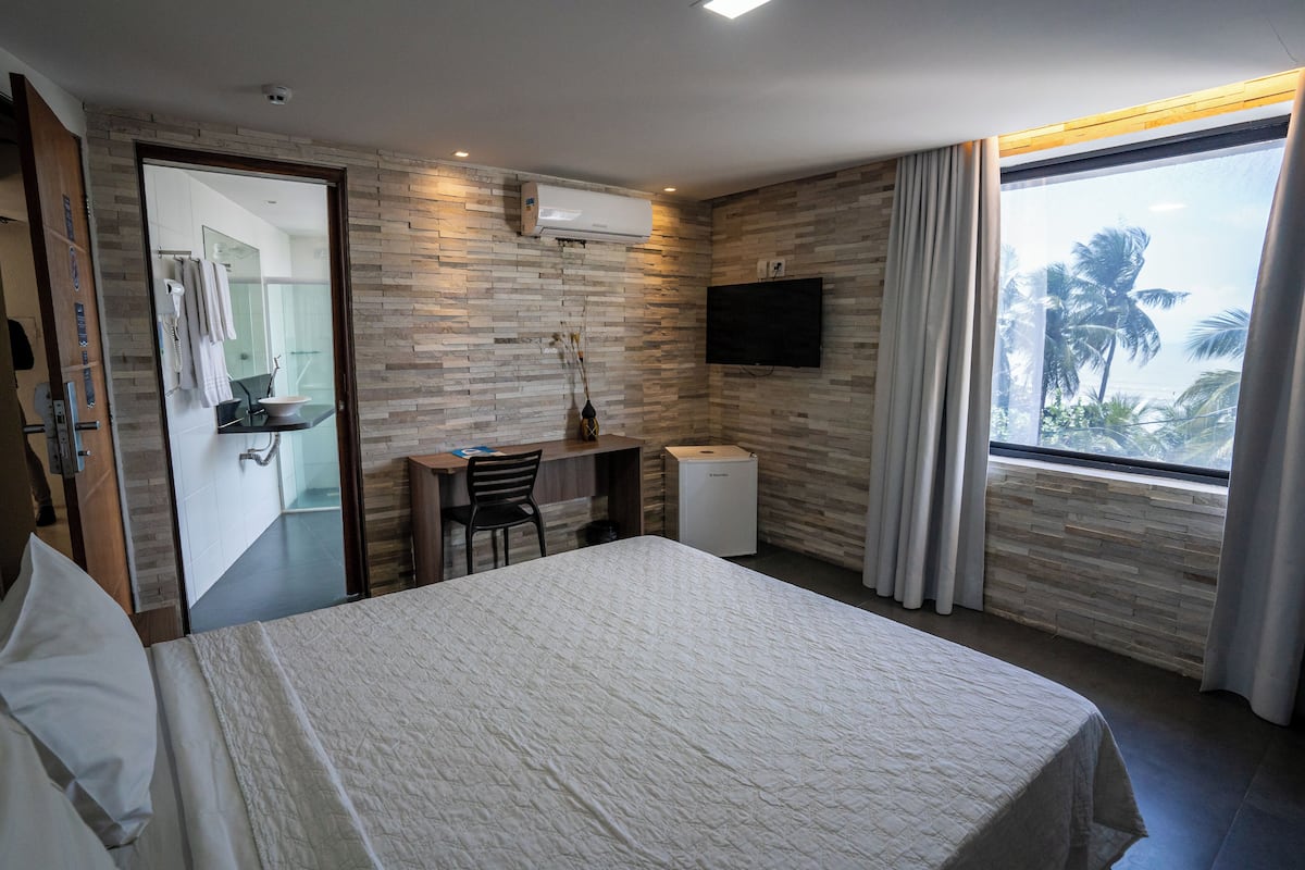 Una camera d'albergo moderna con un letto grande, una scrivania con una sedia, un piccolo frigorifero, una TV a parete e vista su palme attraverso la finestra.