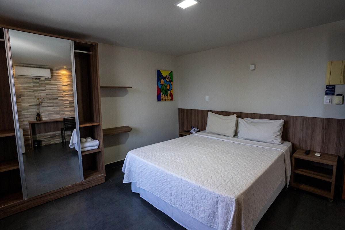 Una camera d'albergo con un letto, un armadio in legno, uno specchio, un tavolino con una sedia e un quadro appeso al muro.