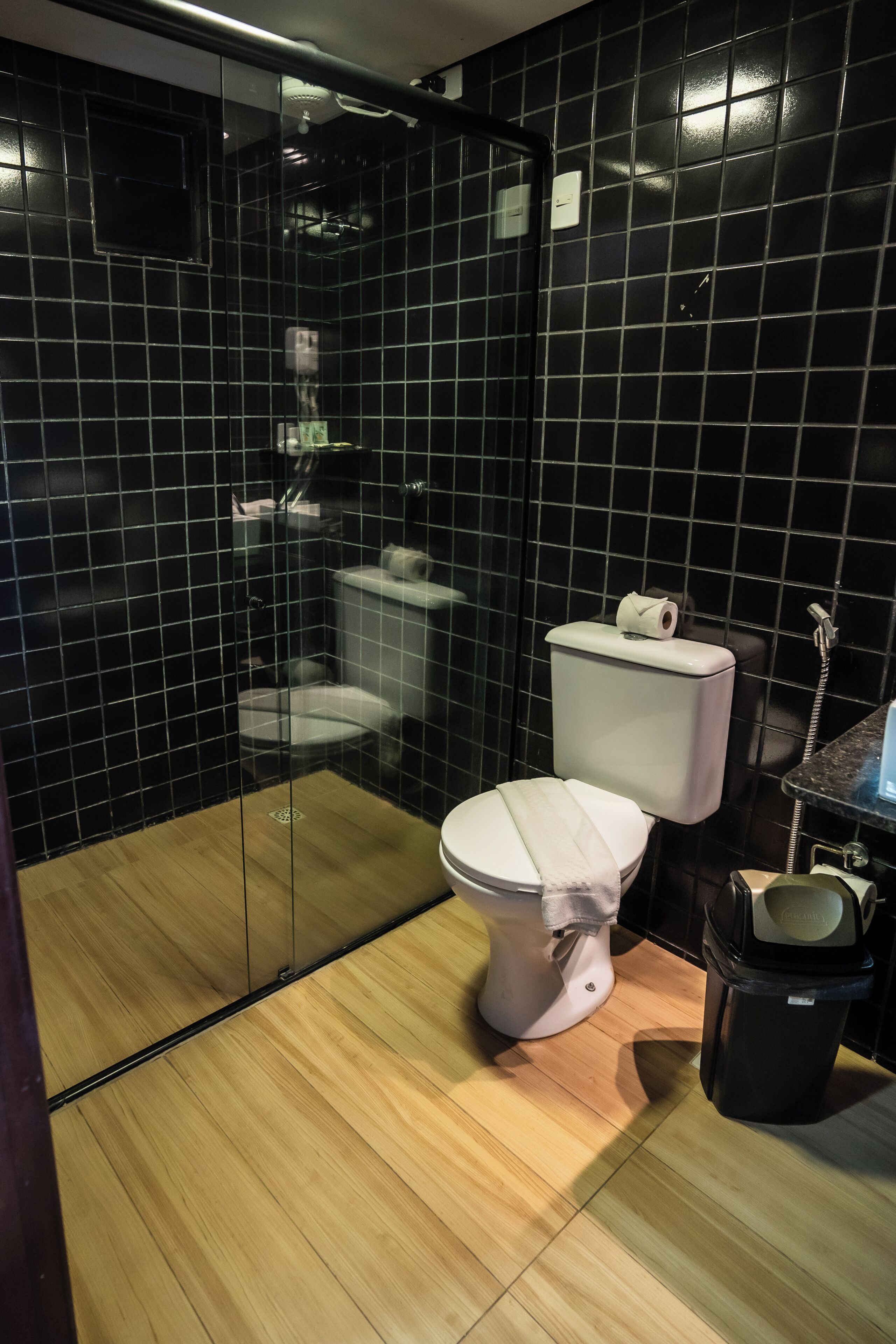 Un baño moderno con paredes de azulejos negros, suelo de madera, inodoro blanco y cabina de ducha con mampara de cristal.