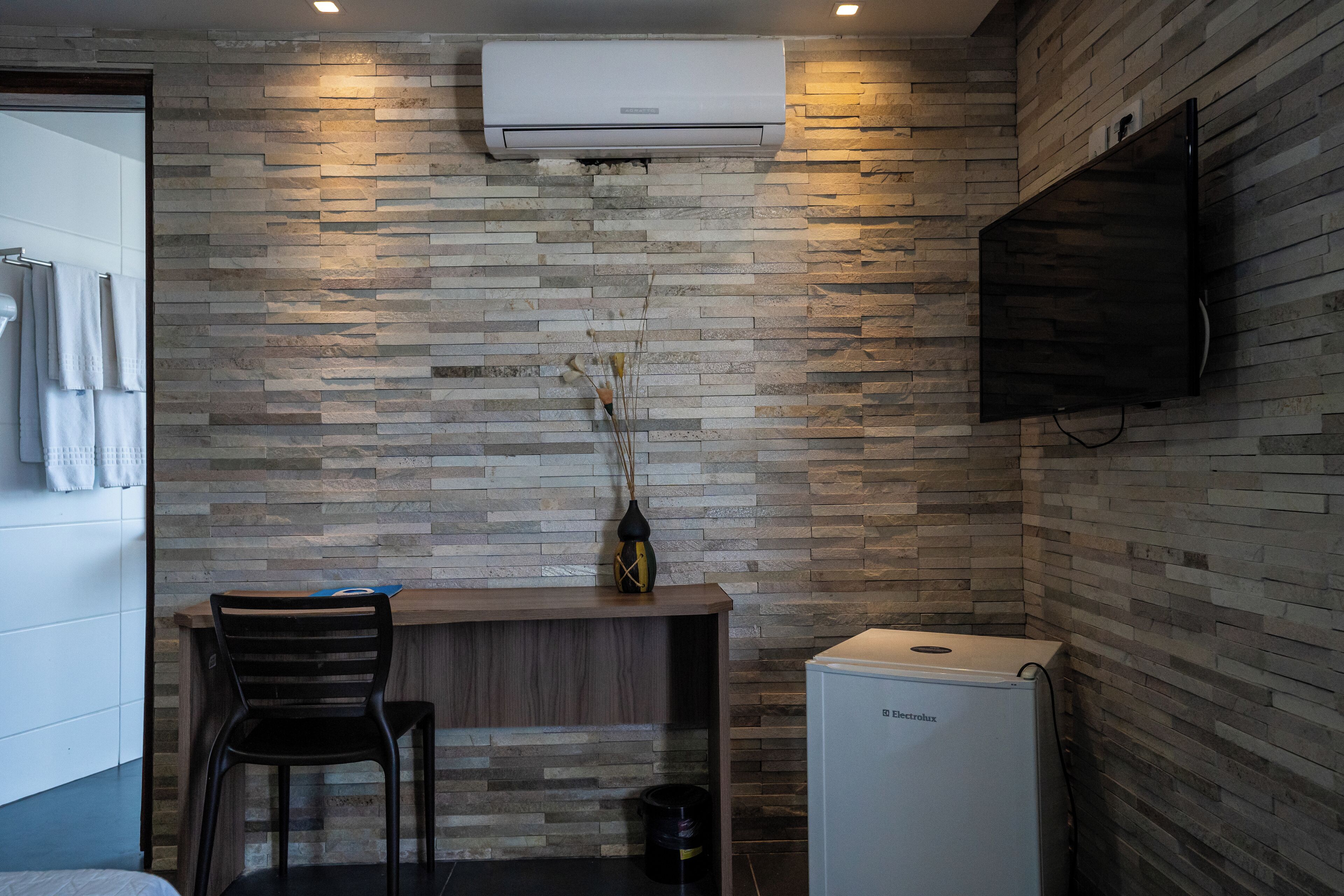 Ein modernes Badezimmer mit Steinwand, einem angebrachten Fernseher, einem kleinen Schreibtisch mit Stuhl und einem kompakten Kühlschrank.