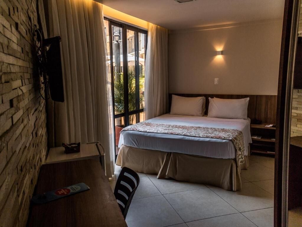 Hotelkamer met een bed, een aan een bakstenen muur bevestigde televisie, een raam met gordijnen en uitzicht op groen buiten.