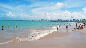 Una playa con gente nadando y relajándose, un horizonte urbano al fondo y un cielo azul despejado.