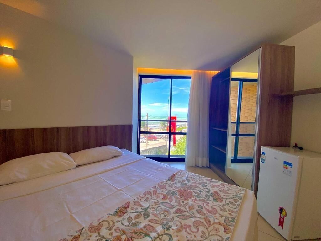 Habitación de hotel con cama, un refrigerador pequeño y vista a la playa a través de un ventanal grande.