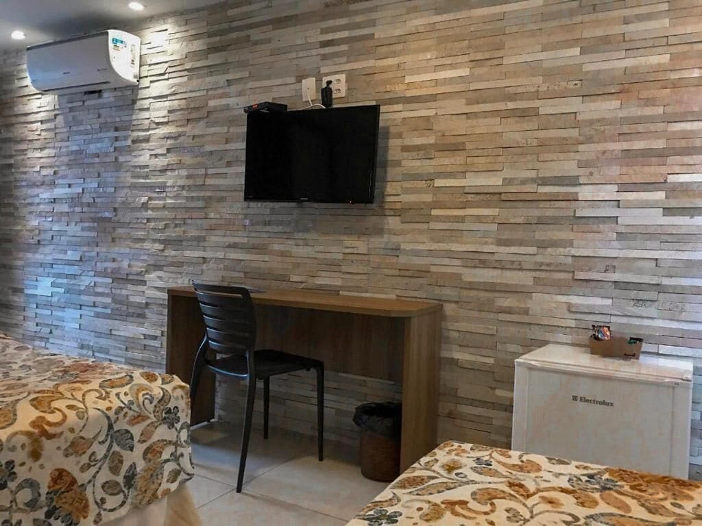 Een kamer met een muur met stenen patroon, een flatscreen televisie boven een houten bureau, een stoel en een klein wit kastje met de tekst 'Electrolux'.