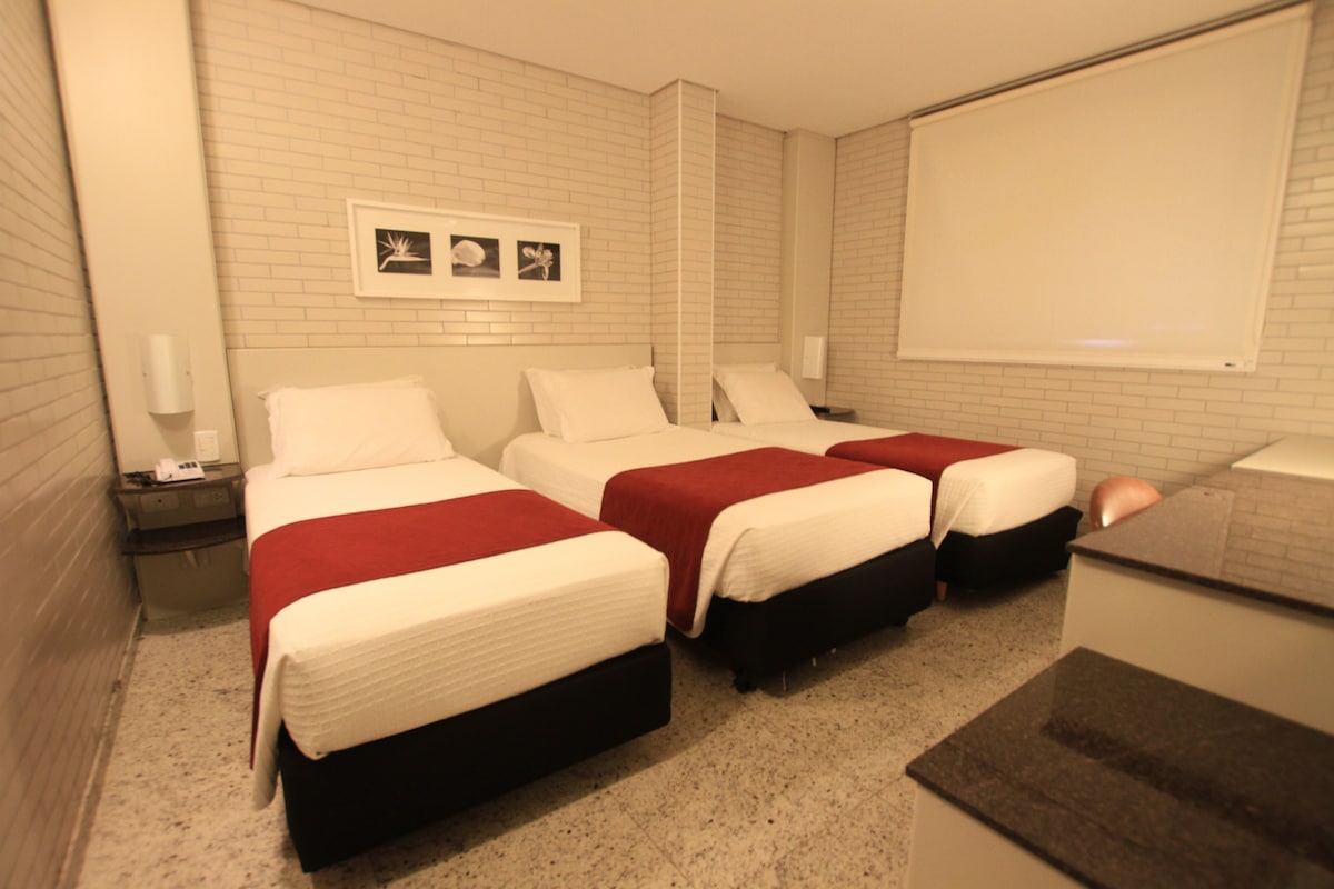 Une chambre d’hôtel avec trois lits, un mur en briques blanches, un écran de projection et un petit bureau avec une lampe.