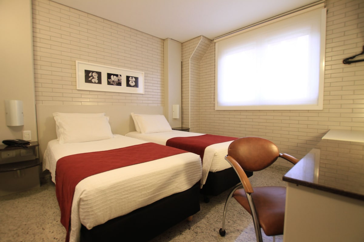 Une chambre d’hôtel avec deux lits, un bureau, une chaise, une fenêtre et un tableau au mur.