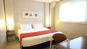 Une chambre d’hôtel avec un lit, une chaise, un bureau et une fenêtre avec des stores.