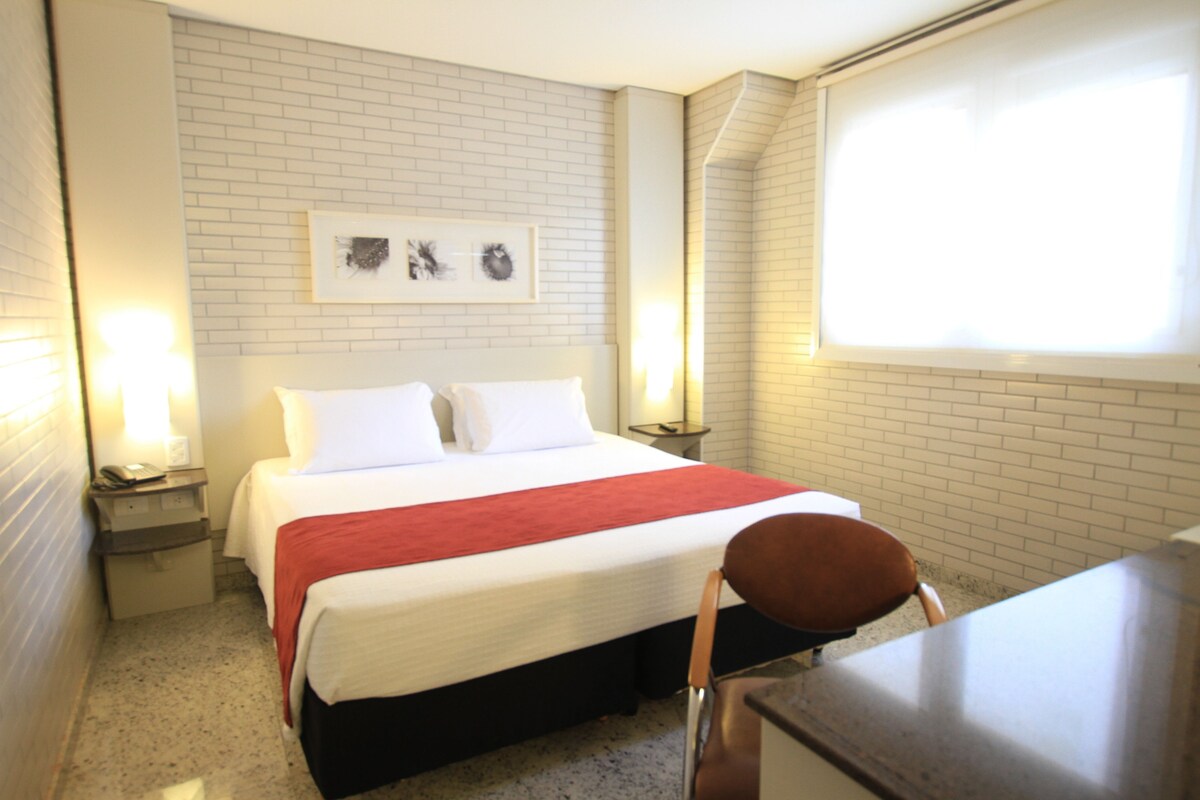 Une chambre d’hôtel avec un lit, une chaise, un bureau et une fenêtre avec des stores.