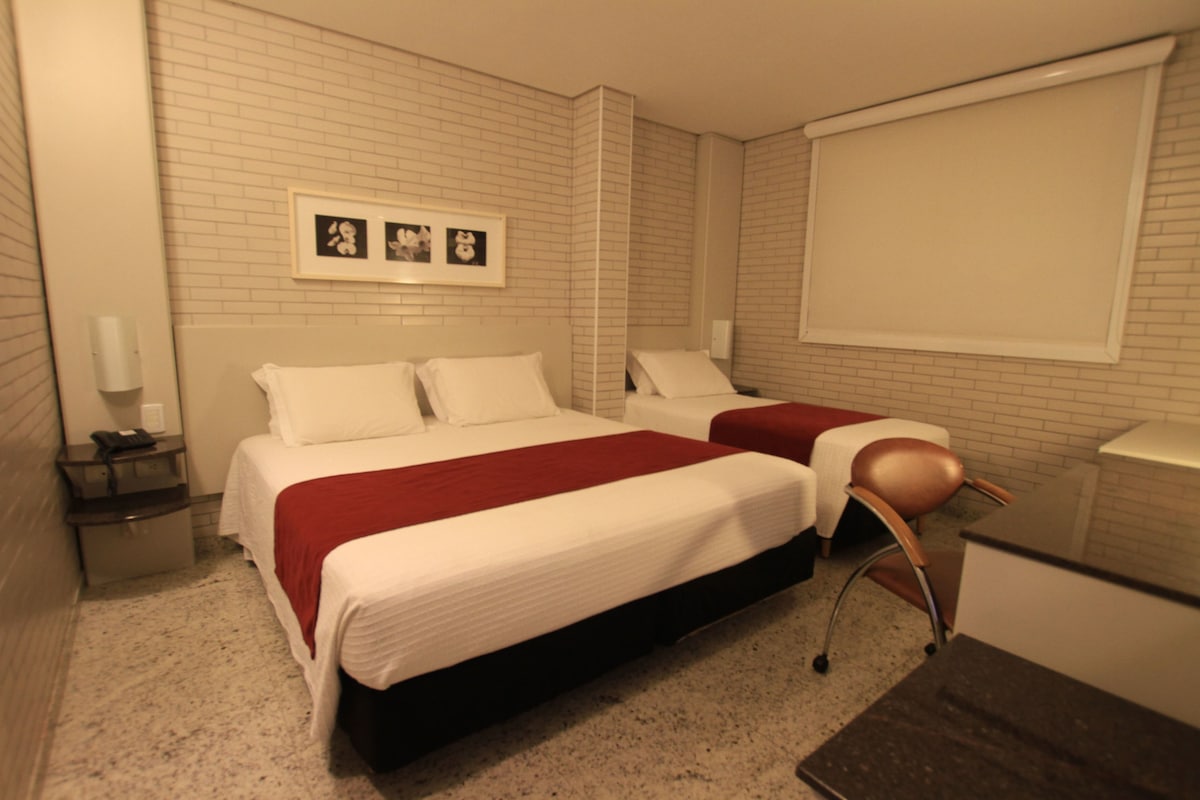 Une chambre d’hôtel avec un grand lit, un lit plus petit, un bureau et une chaise. Il y a un tableau au mur et une fenêtre avec un store.