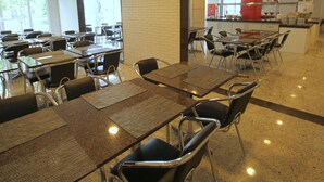 Une cafétéria avec des tables et des chaises, un espace cuisine doté d’un comptoir rouge, et une vue sur l’extérieur grâce à de grandes fenêtres.