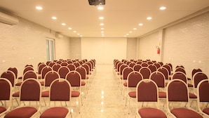 Un salón de conferencias con hileras de sillas rojas y mesas blancas, unidades de aire acondicionado en las paredes y un extintor en la pared derecha.