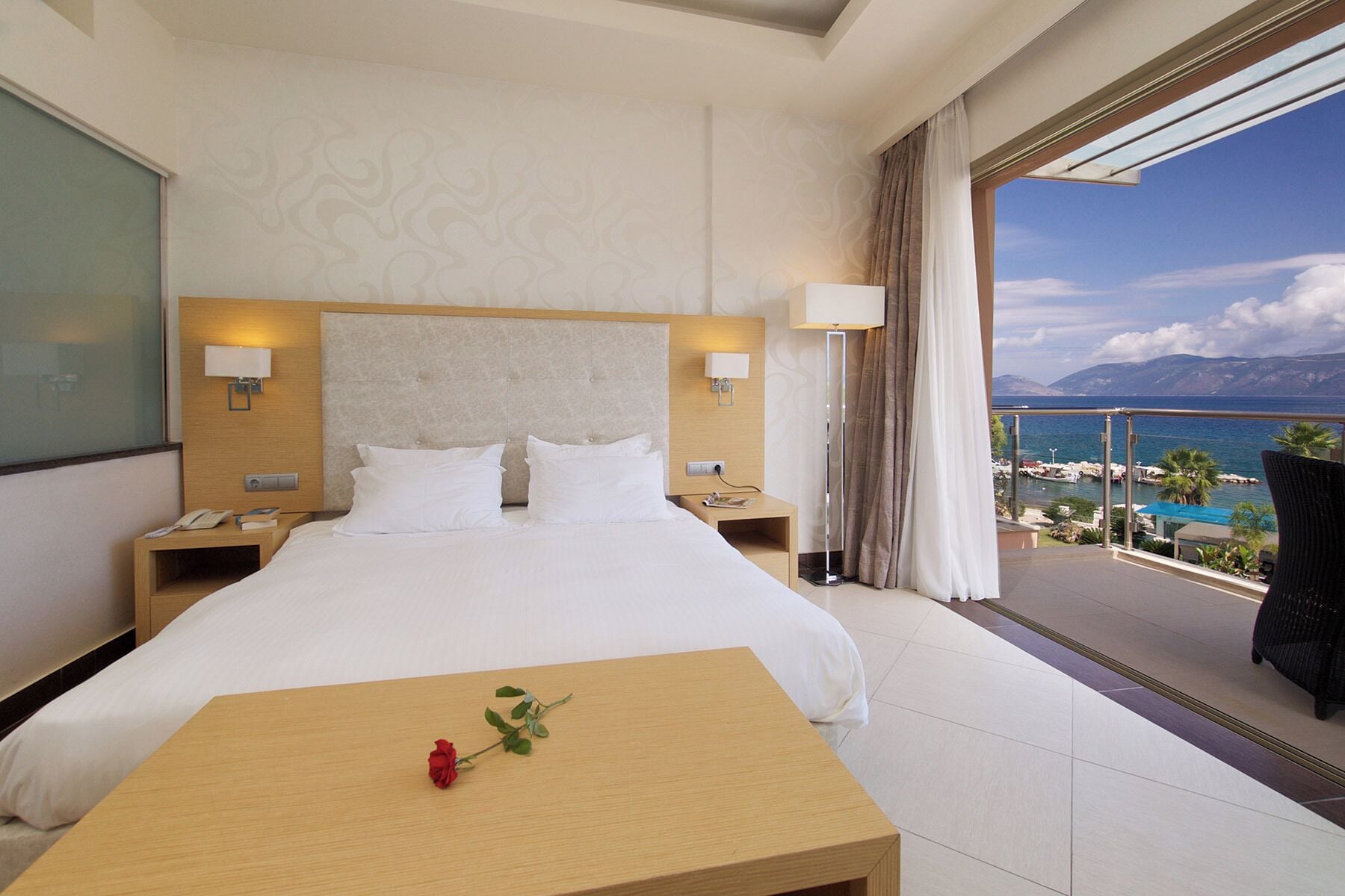 A hotel room with a bed, bedside tables, a nightstand, a small table with a rose, and a balcony with a view of the sea and boats.