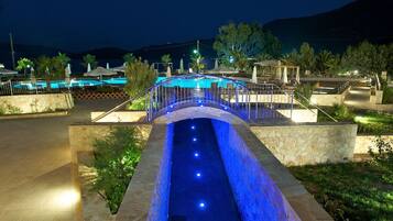 Relax by the serene pool, surrounded by lush greenery and palm trees, with a gentle bridge and lounge chairs nearby.