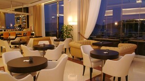 En moderne hotellobby med runde borde og sofaer, et barområde og store vinduer med gardiner.
