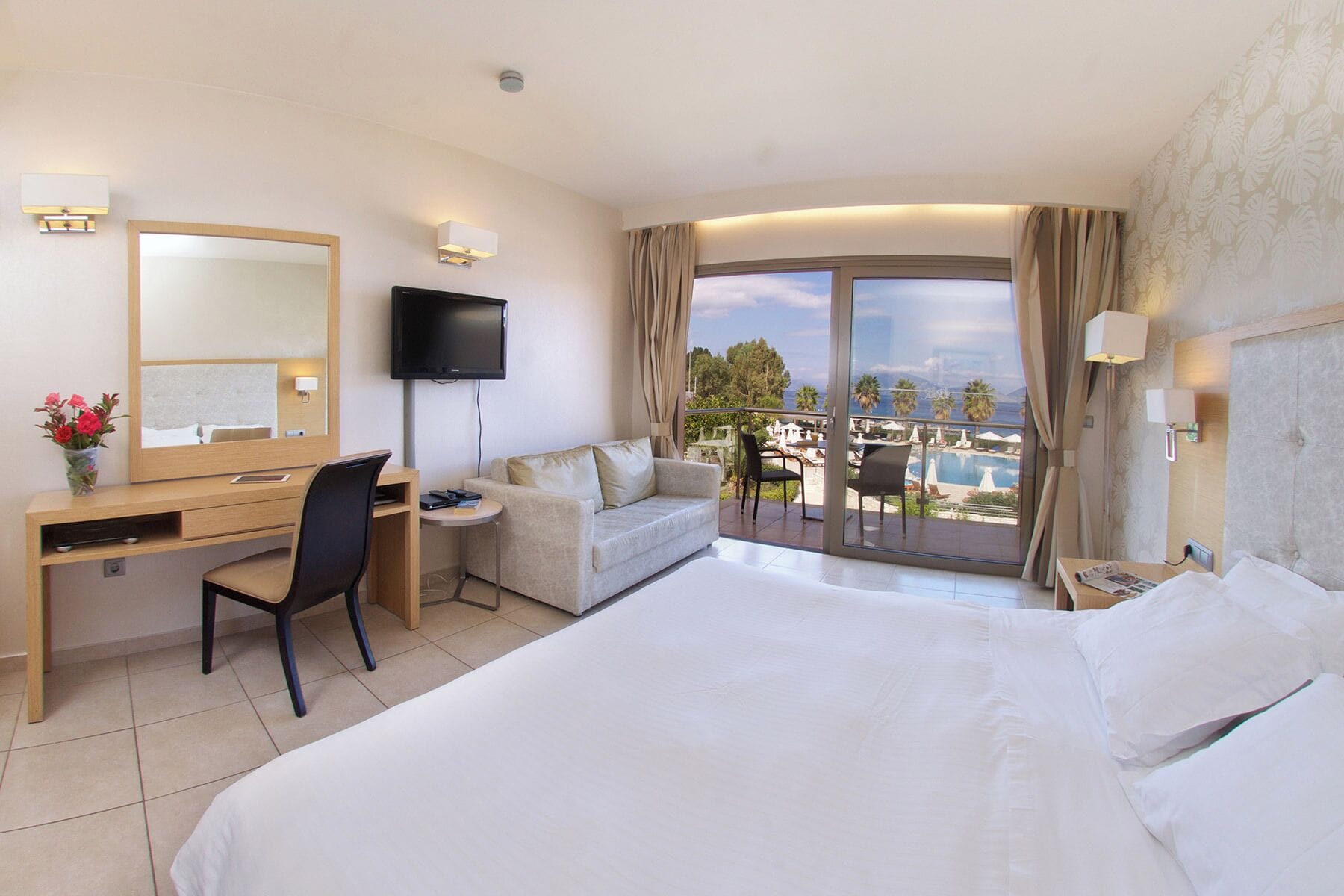 A hotel room with a bed, desk, chair, TV, and a balcony with a pool view.