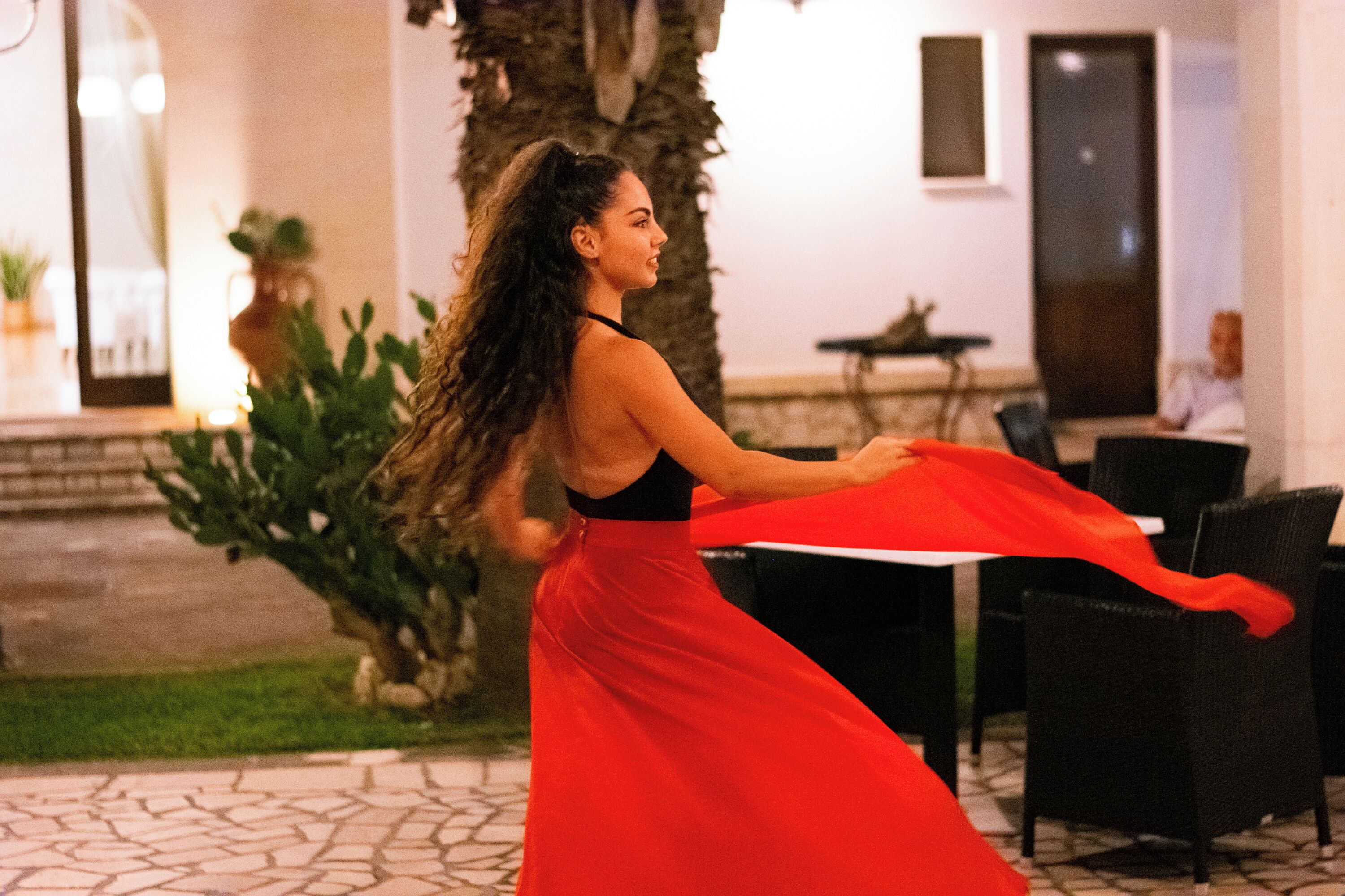 A woman in a red dress dancing outdoors, with a palm tree and a building in the background.
