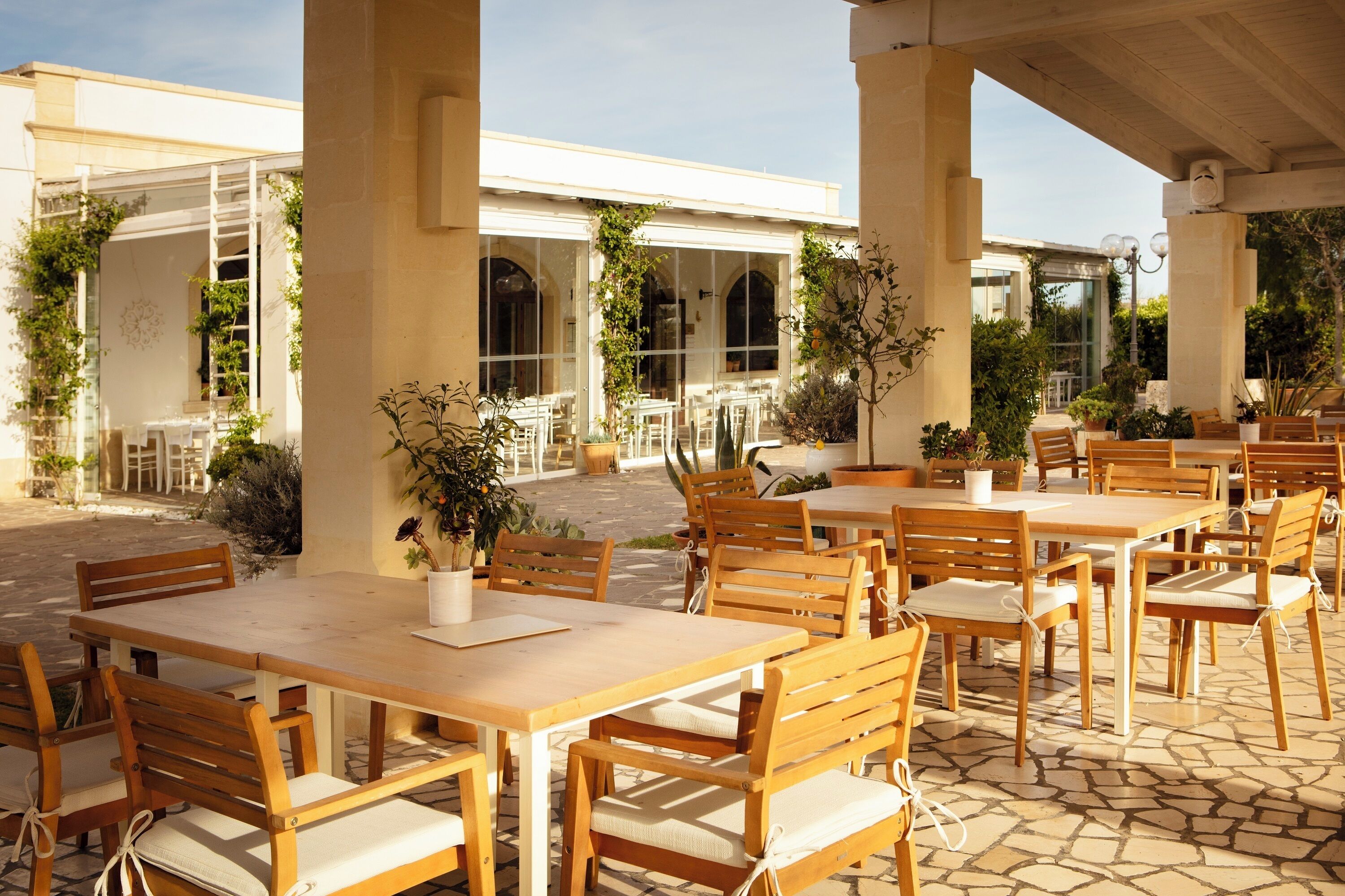 Un espacio al aire libre con un patio que tiene varias mesas y sillas de madera, rodeado de columnas grandes y plantas en macetas.