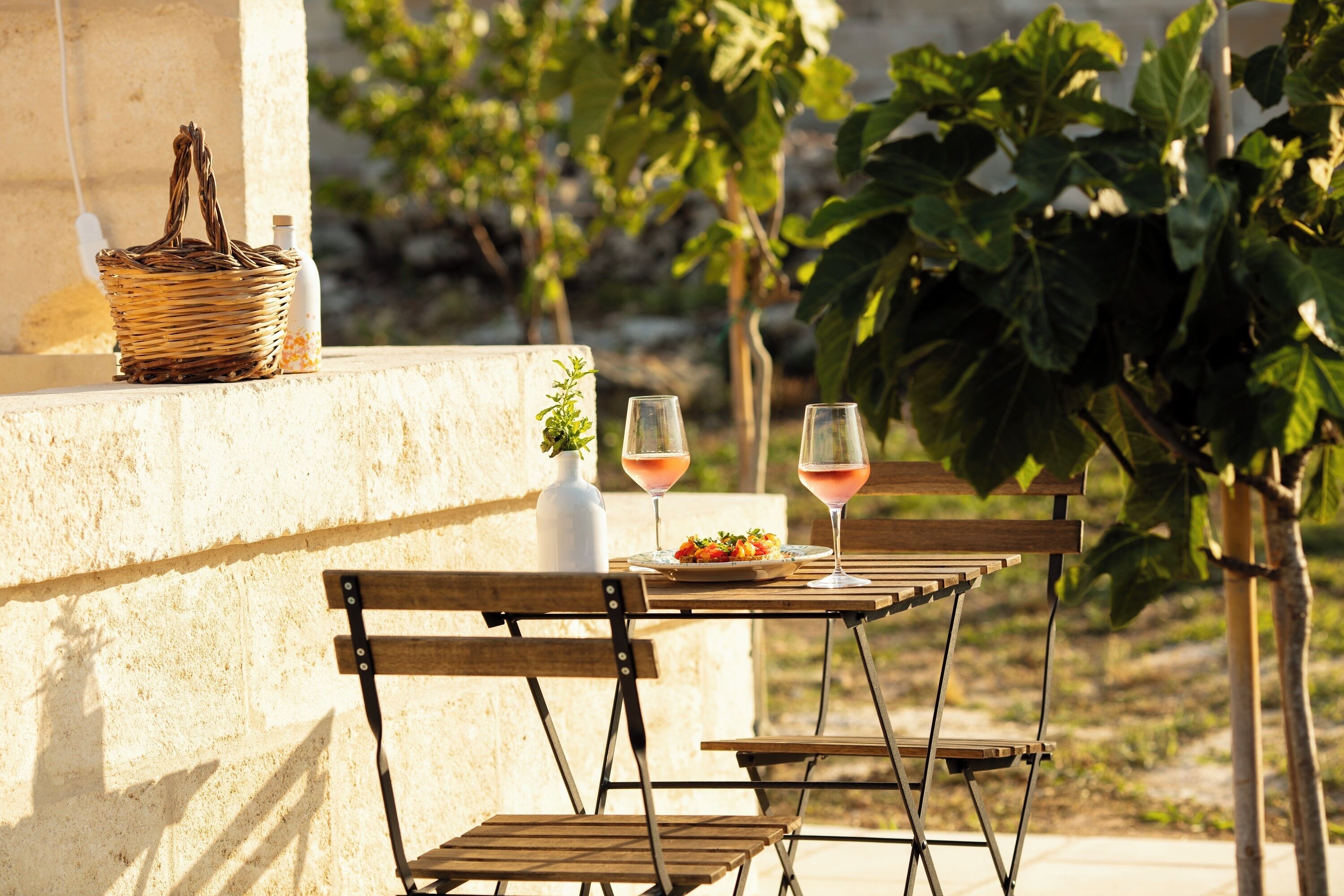 Un espacio de comedor al aire libre con una mesa, sillas, una cesta de mimbre, un jarrón blanco y dos vasos de bebida rosada.