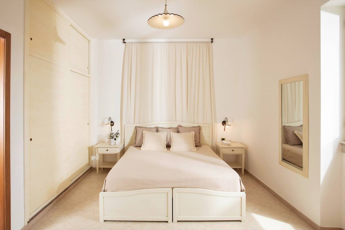 A bedroom with a double bed, two bedside tables, a nightstand, a mirror, and a ceiling light fixture.