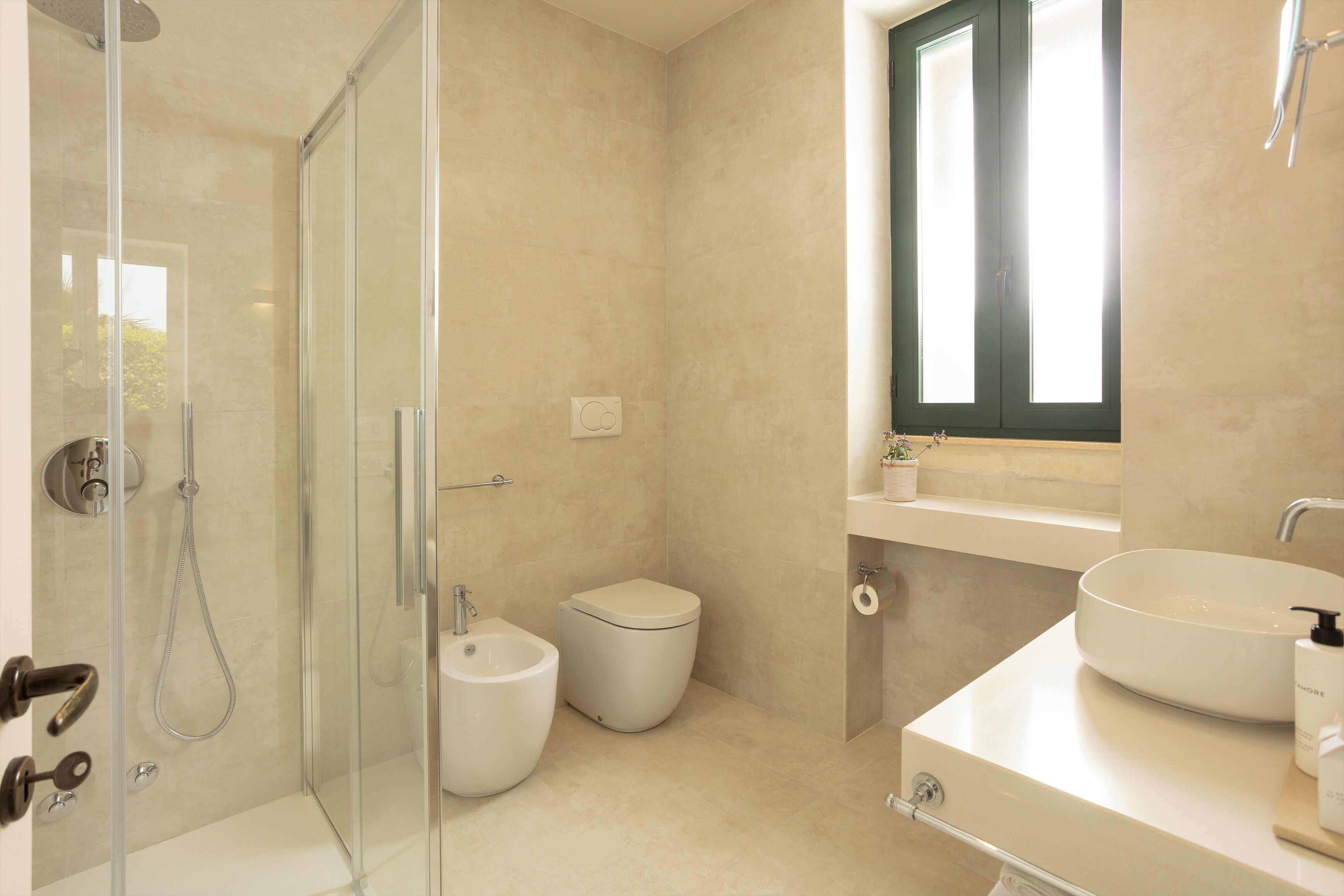 Une salle de bain moderne dotée d’une douche à parois de verre, d’un bidet, de toilettes et d’un lavabo placé sous une fenêtre.