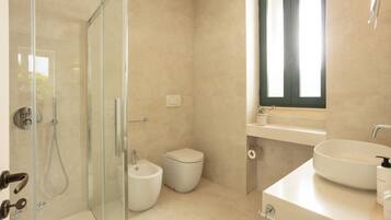 Une salle de bain moderne dotée d’une douche à parois de verre, d’un bidet, de toilettes et d’un lavabo placé sous une fenêtre.