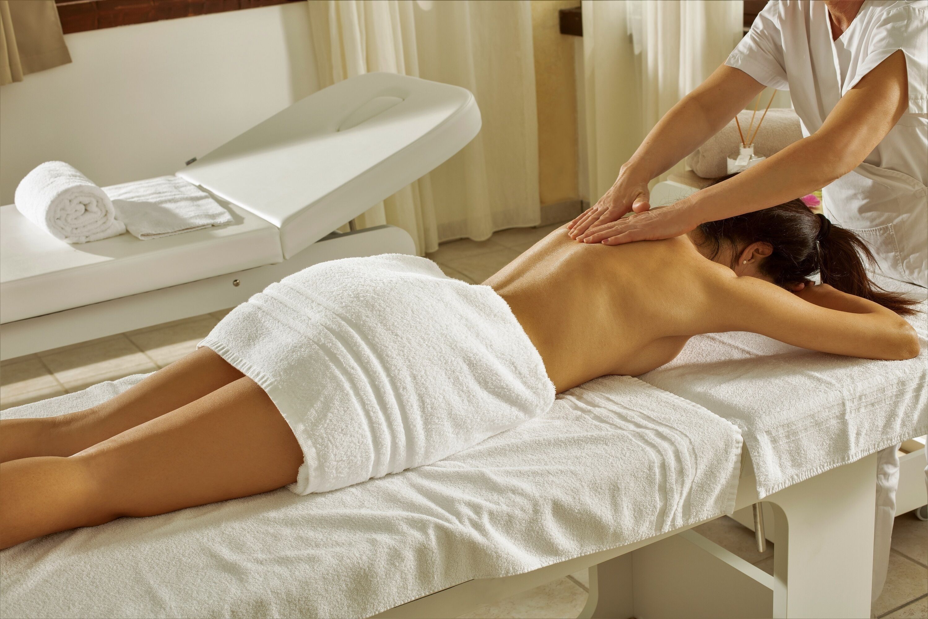 A massage therapy session in a clinical setting with a client lying on a treatment table, a therapist attending to the client, and a rolled towel on a side table.