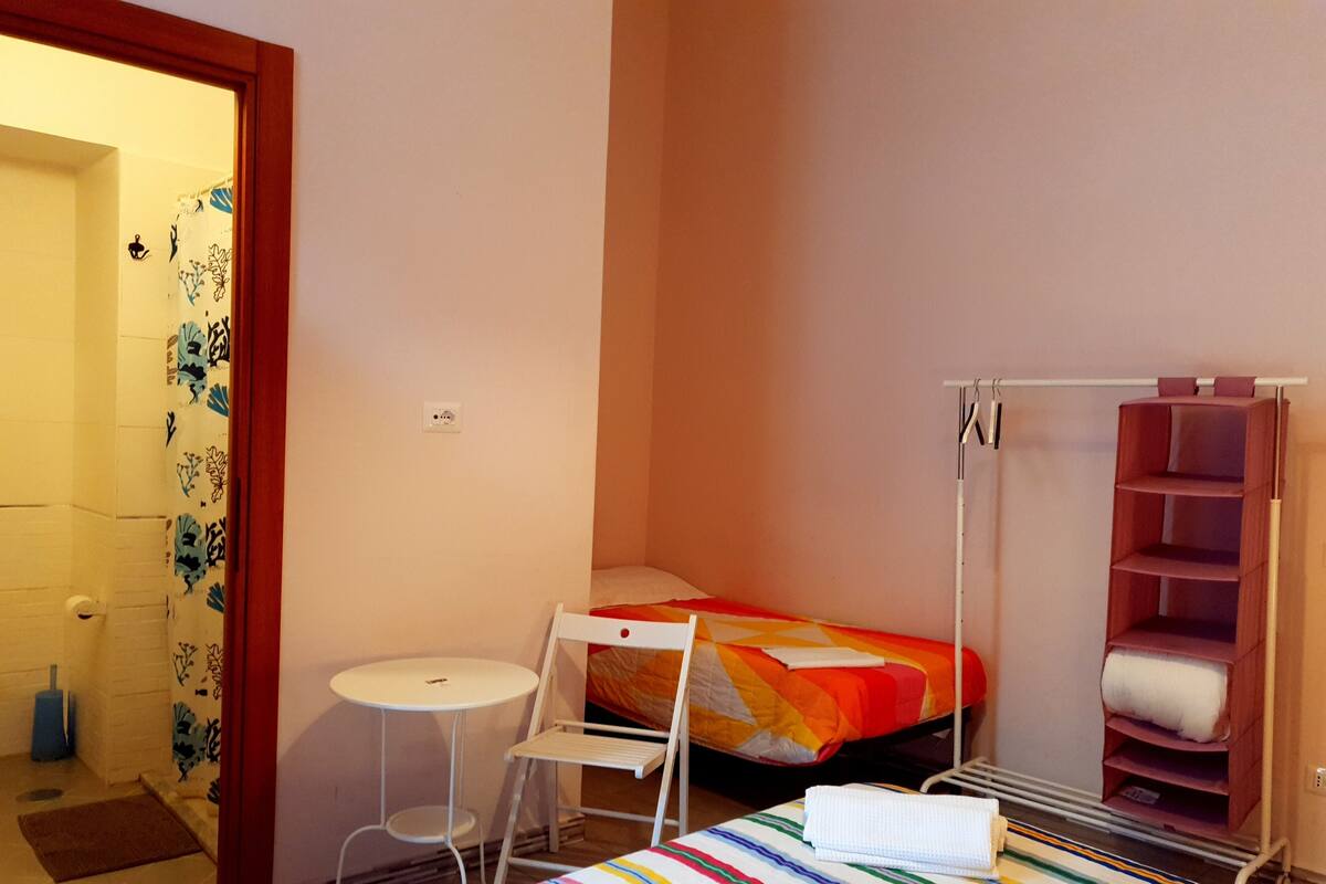 A small, neatly arranged bedroom with a bed, a small table, a chair, and a wardrobe.