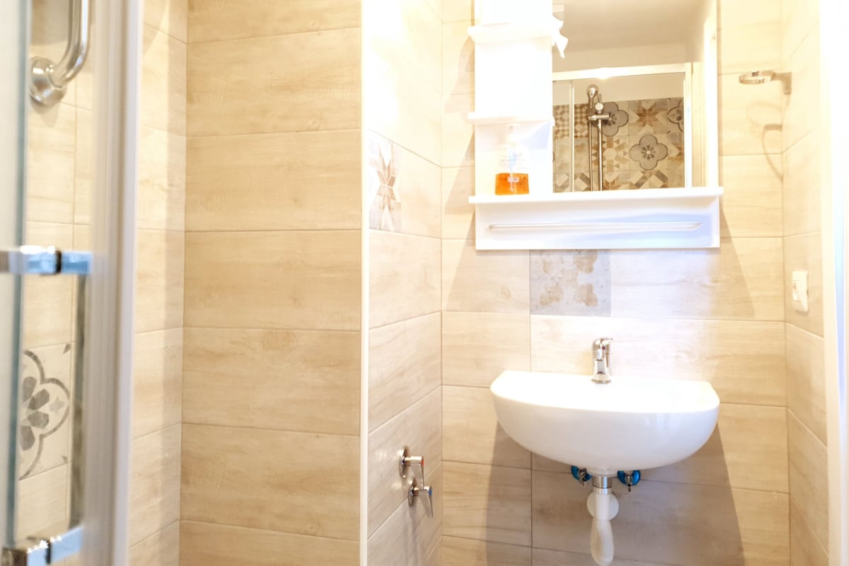 Un baño con baldosas color beige, un lavamanos blanco con un espejo encima y una zona de ducha visible a través de una puerta abierta.