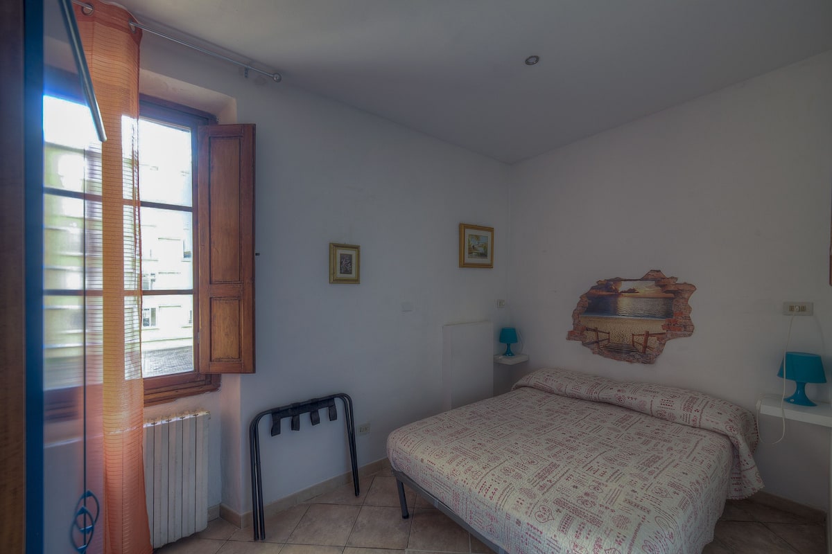 A bedroom with a bed, a window with curtains, a radiator, a wall-mounted shelf with a lamp, and a picture frame.