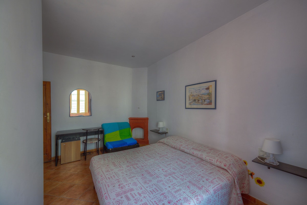 A bedroom with a bed, a desk, a chair, and a small table with a lamp.