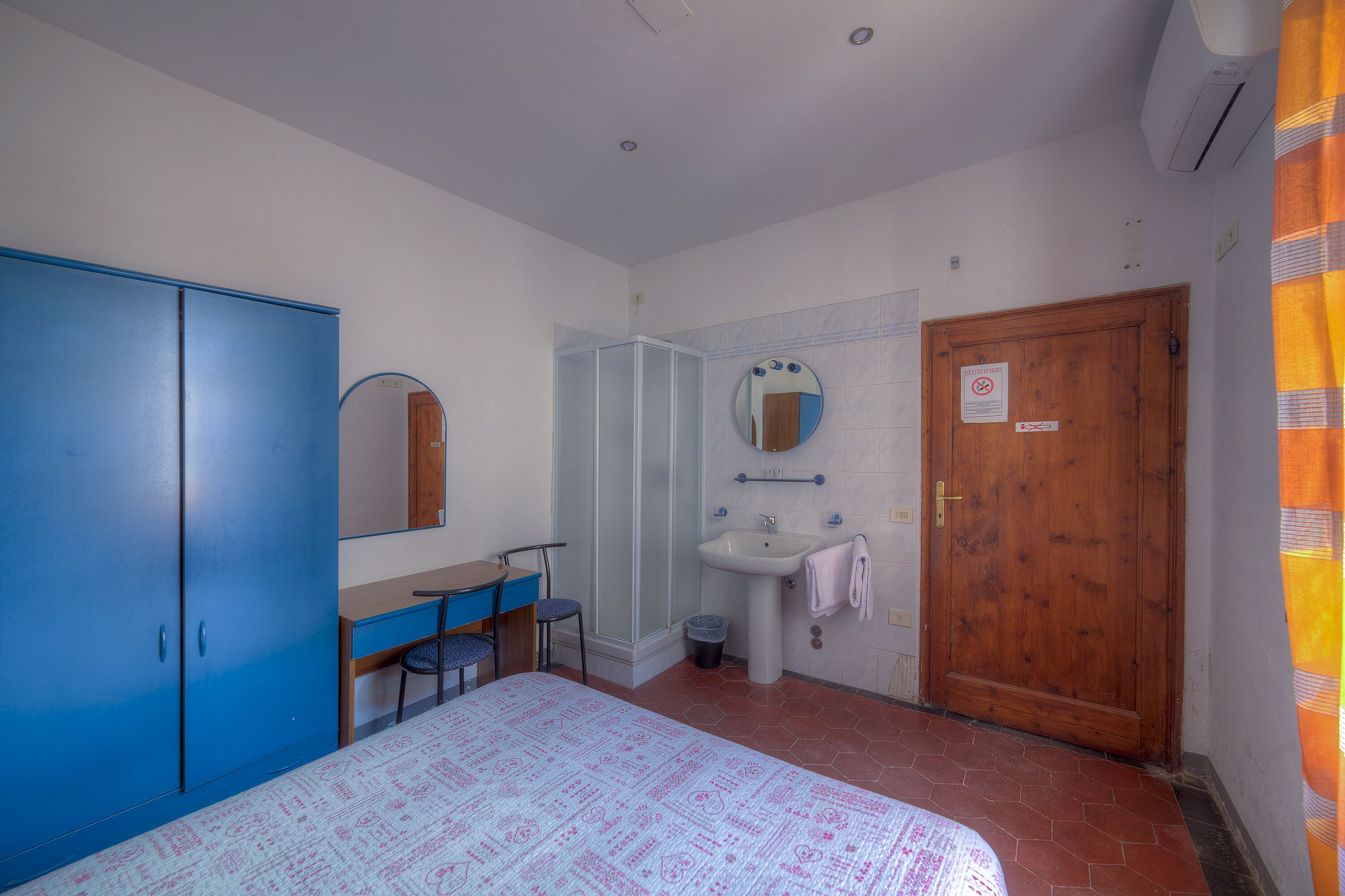 A bedroom with a bed, a desk, a chair, a wardrobe, a mirror, a sink, and a shower.