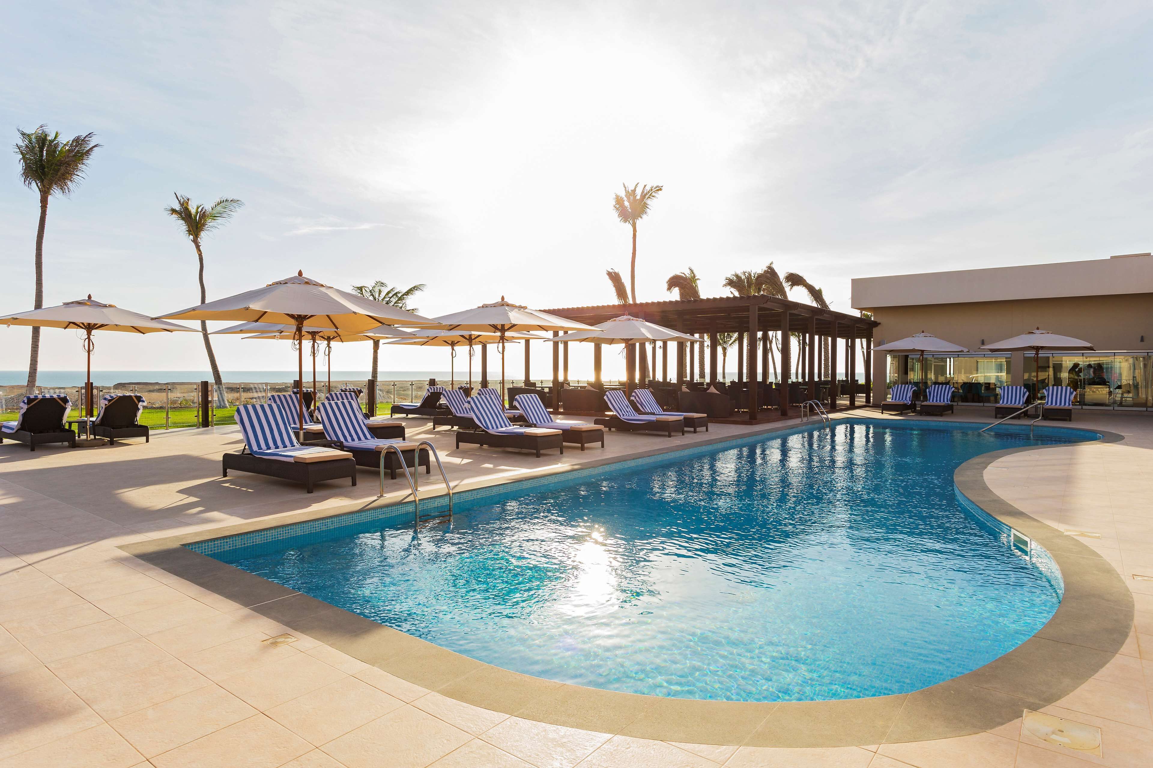 A pool area with lounge chairs and umbrellas, a clear sky, and palm trees.