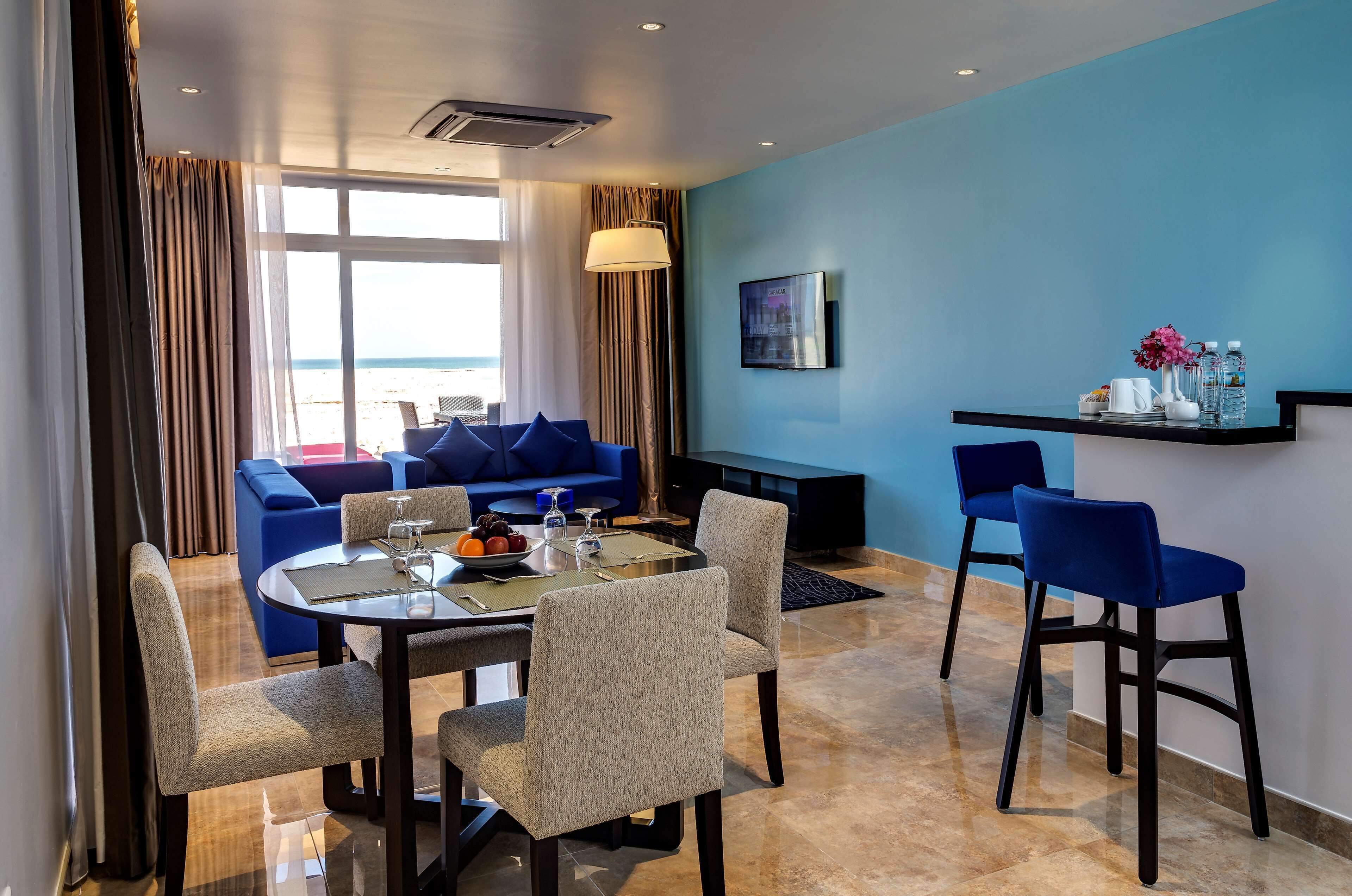 Una habitación de hotel moderna con zona de comedor, un conjunto de sofás y un televisor montado en la pared.