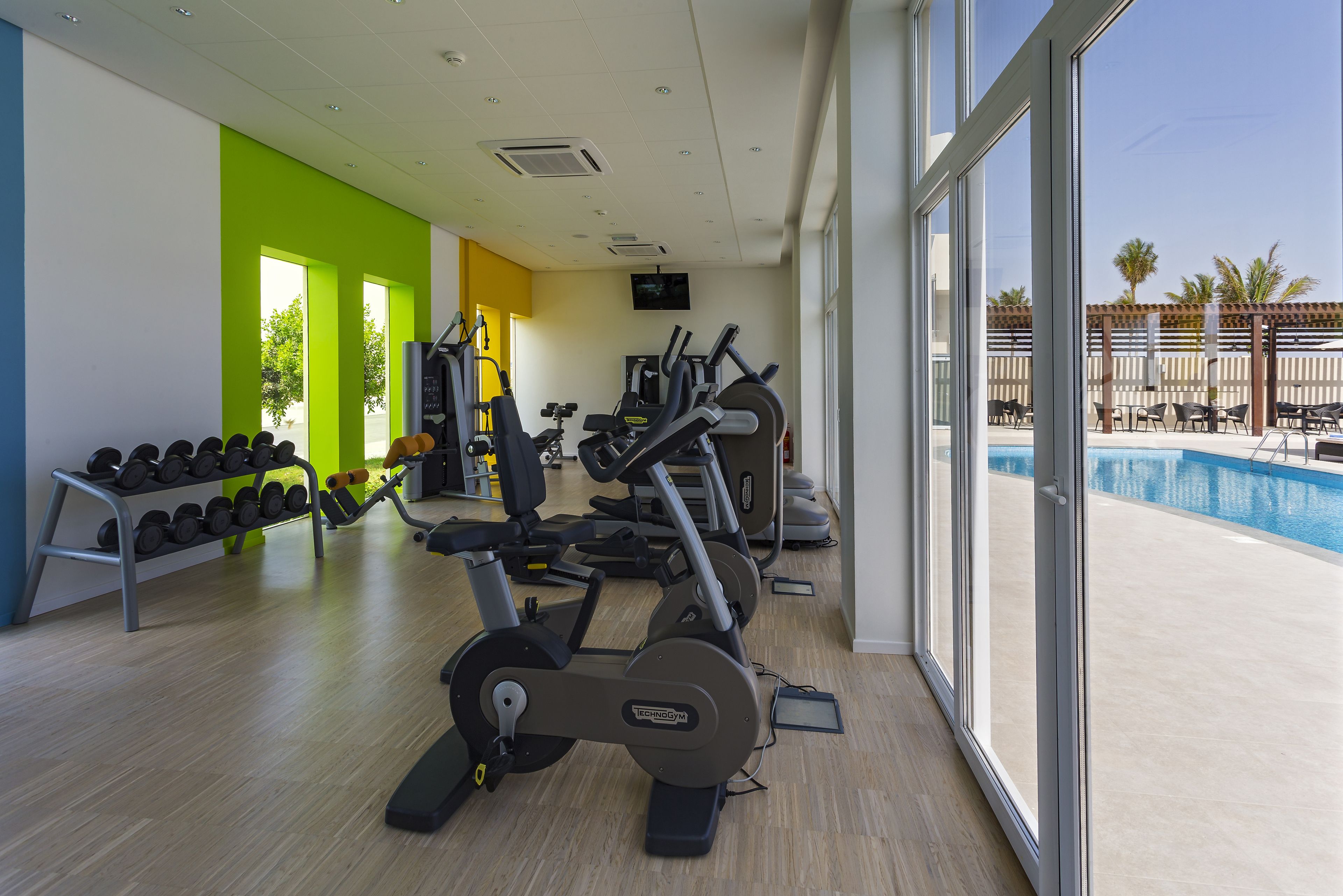 A gym with cardio machines, dumbbells, and a pool area visible through large windows.