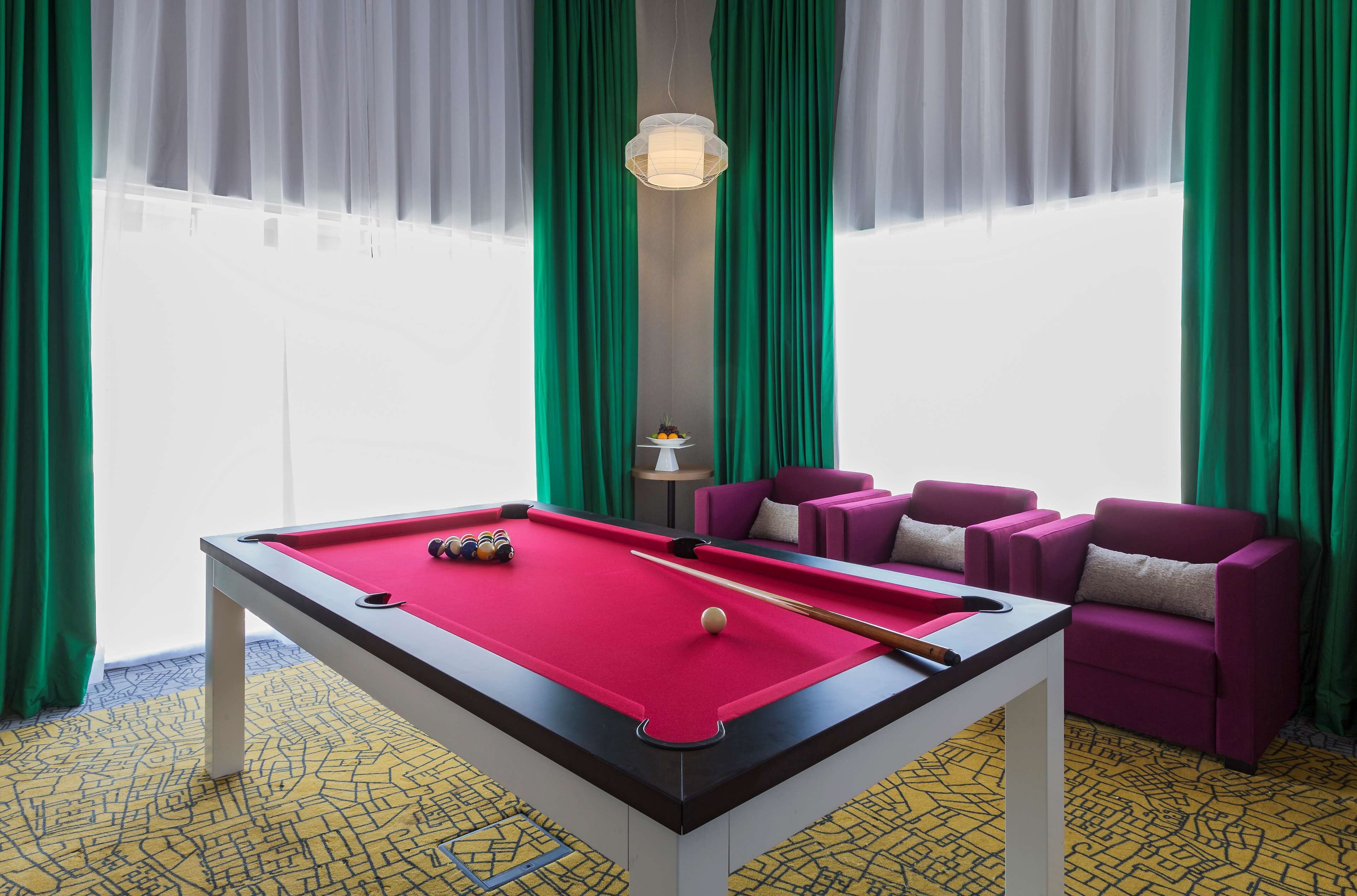 A pool table with a red felt surface, surrounded by purple chairs and a small table with a lamp.
