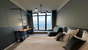 A hotel room with a large bed, a desk, a chair, and a view of the sea through the windows. -  L'Echappée - Hôtel Casino Dieppe (Dieppe)