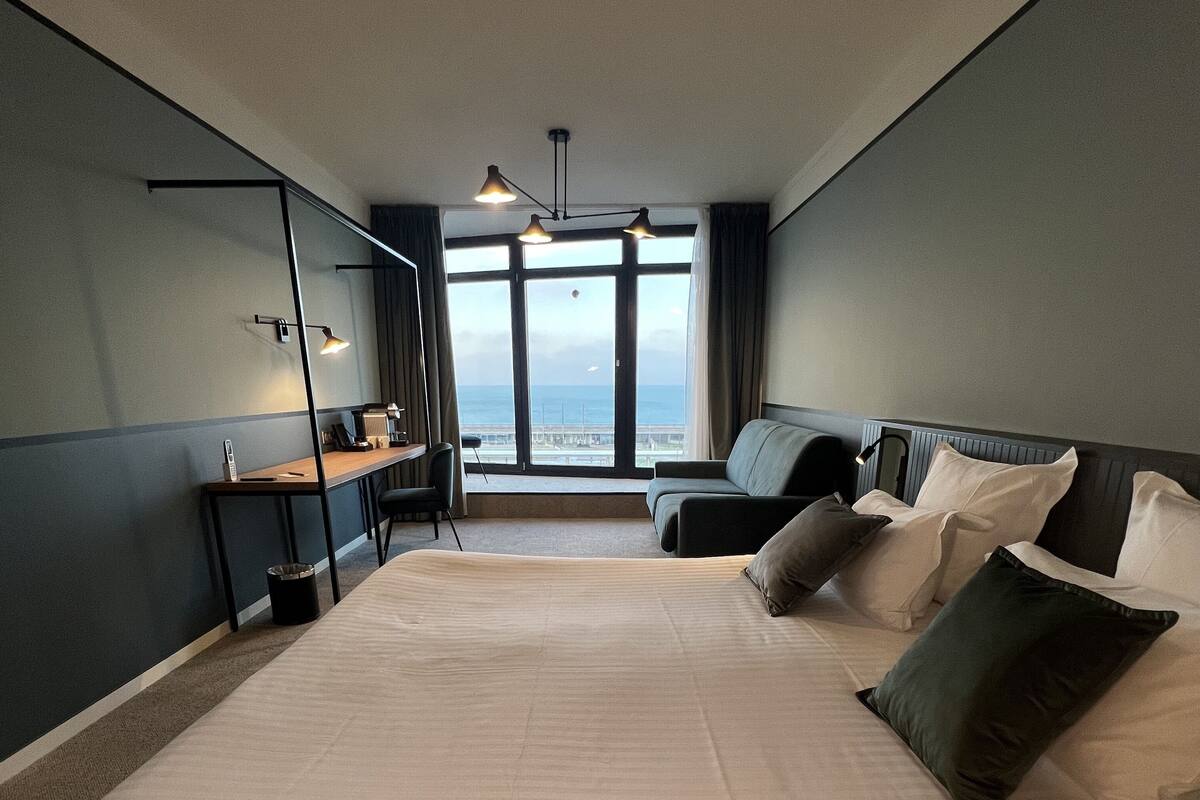 Hotellihuone, jossa on suuri sänky, työpöytä, tuoli ja ikkunoista avautuva näkymä merelle.
