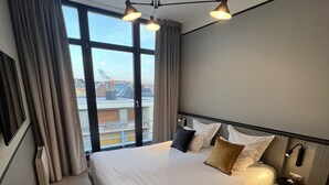 A hotel room with a large bed, a desk, a chair, a nightstand with a lamp, and a view of the city through large windows. -  L'Echappée - Hôtel Casino Dieppe (Dieppe)