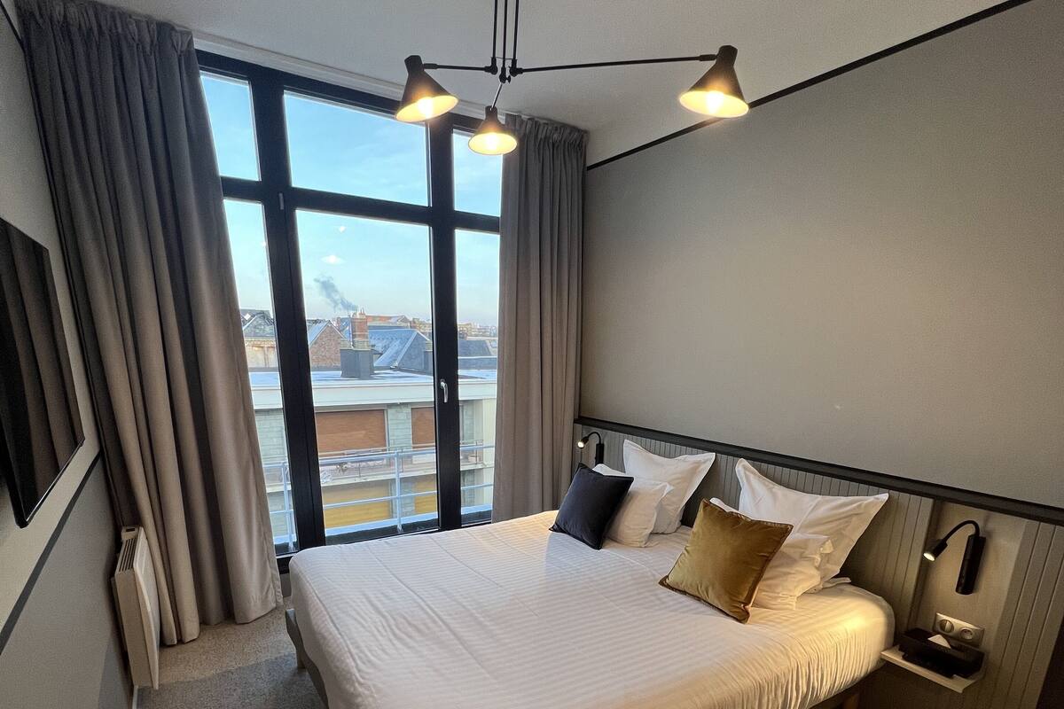 Hotellihuone, jossa on suuri sänky, työpöytä, tuoli, yöpöytä lampun kanssa ja suuret ikkunat, joista avautuu näkymä kaupunkiin.