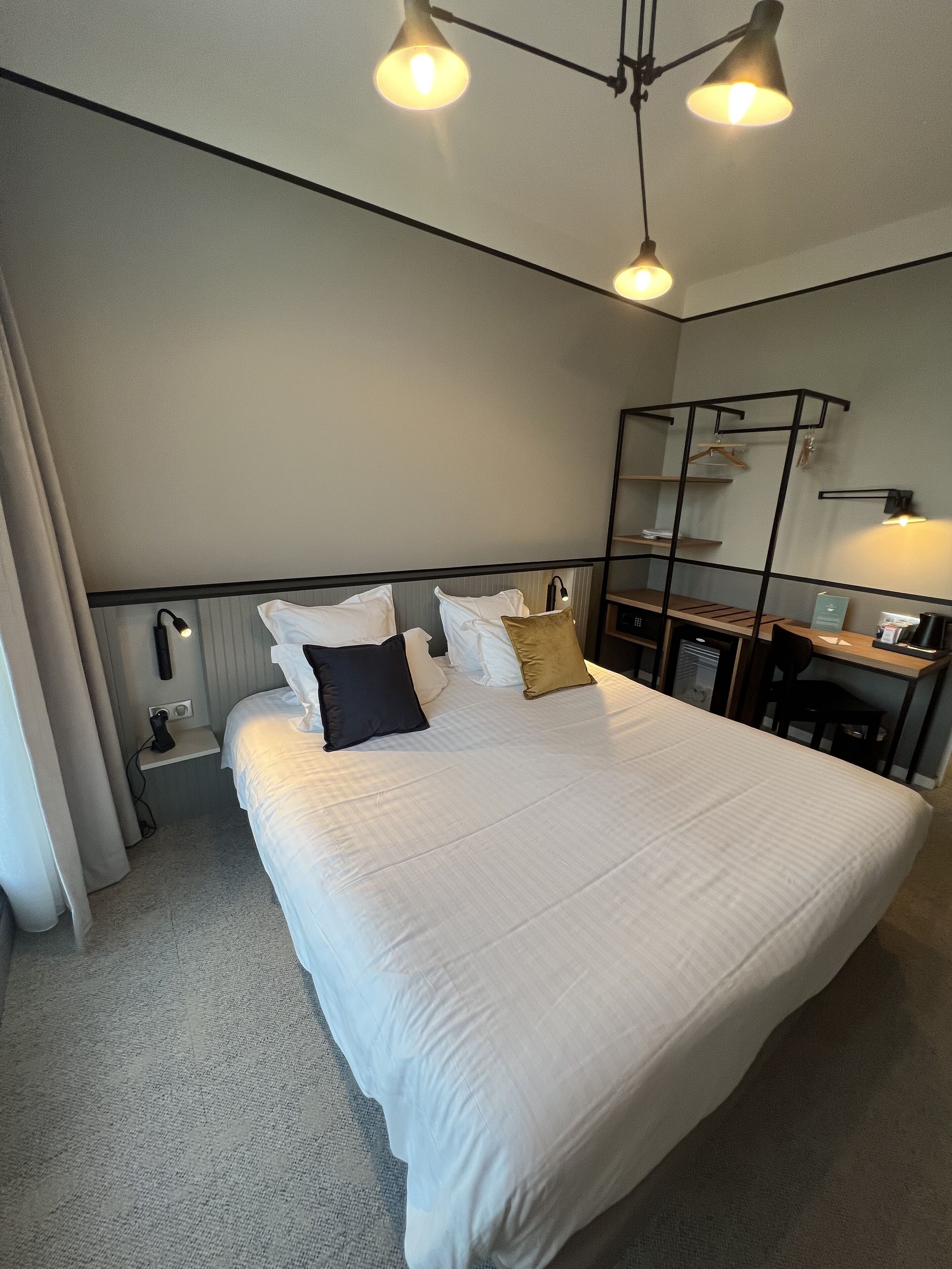 A neatly made bed with white linens and multiple pillows, a bedside table with a lamp, a small desk with a chair, and a mirror on the wall.
