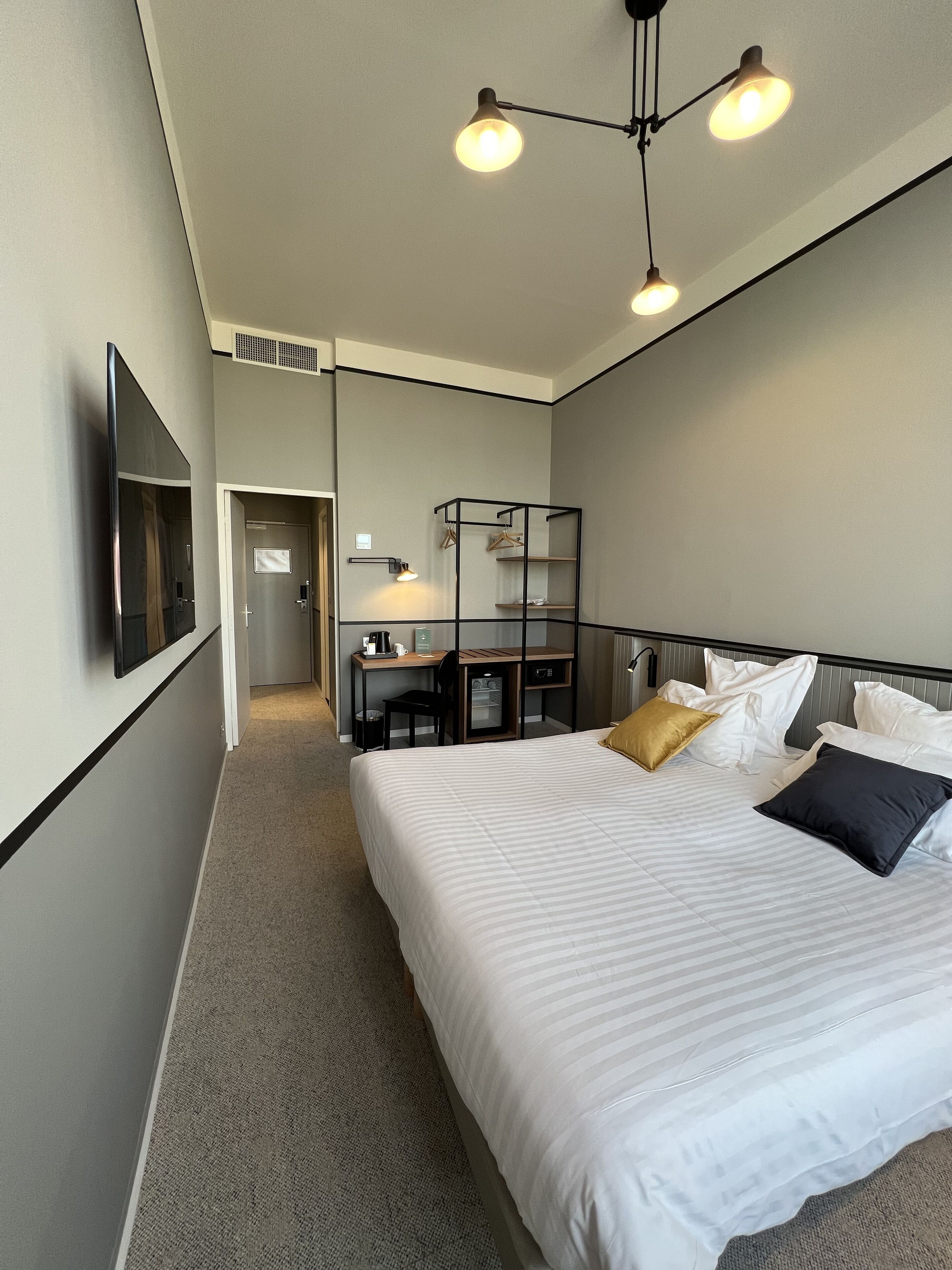 A hotel room with a large bed, a desk, and a chair. There is a television mounted on the wall and a wardrobe.