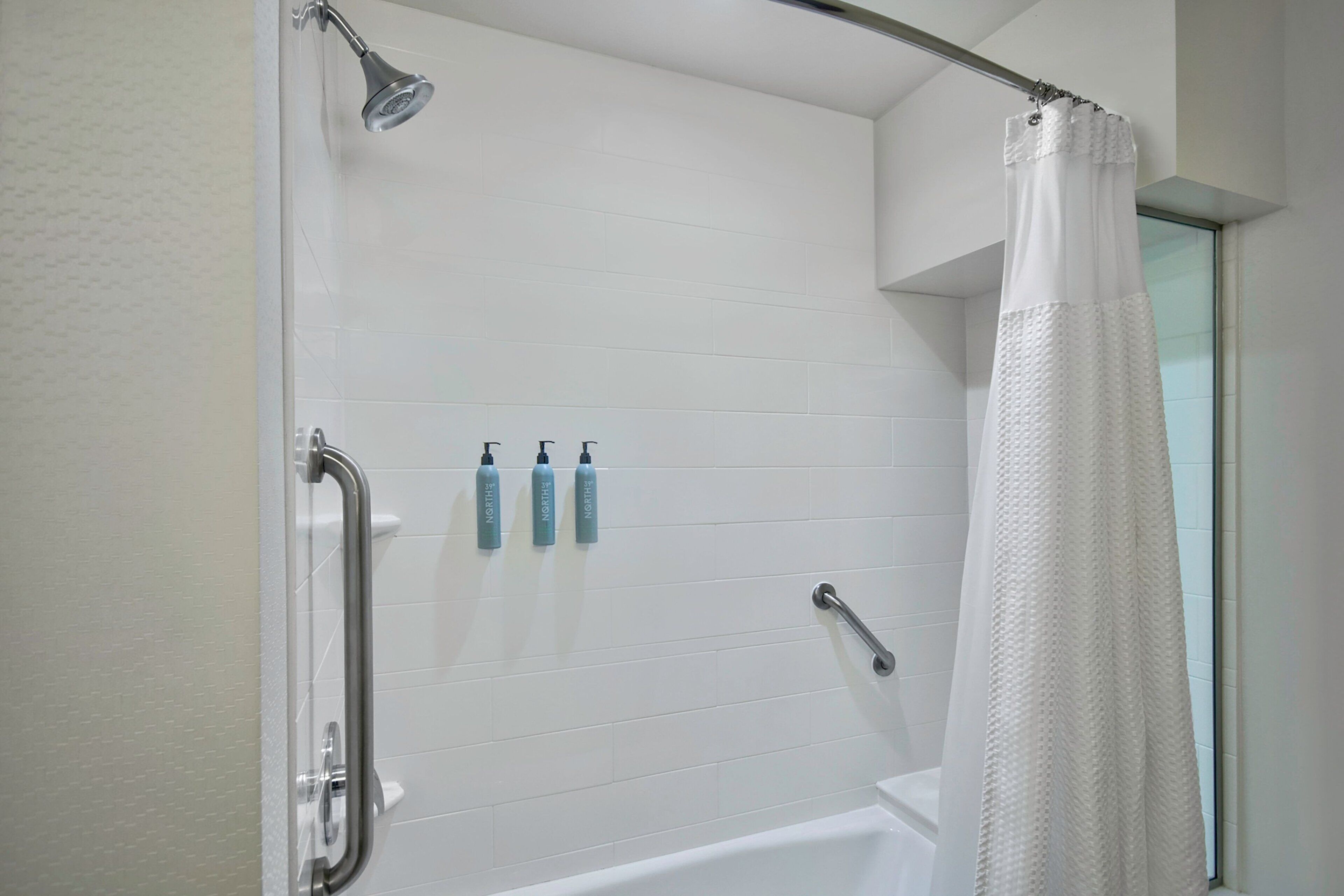 Un espace douche avec un mur recouvert de carreaux blancs, une porte en verre, un rideau de douche blanc et trois bouteilles de shampoing posées sur le mur.